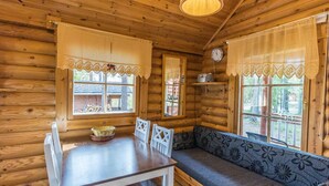 L’intérieur d’un chalet en bois, avec une table à manger, des chaises, un banc, des rideaux en dentelle et une horloge au mur.