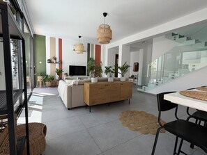 Een moderne woonkamer met een bank, een houten kast en een eettafel met stoelen. Er staan potplanten en een televisie op een dressoir.