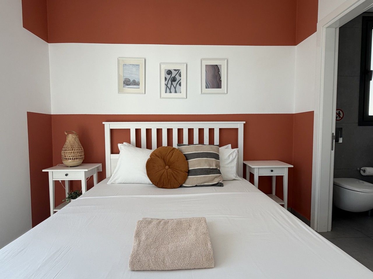 A bedroom with a bed, two bedside tables, a nightstand, a wall-mounted shelf, and a door leading to a bathroom.