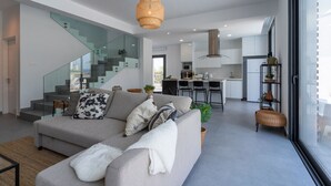 Ein zeitgemäßer Wohnraum mit grauem Sofa, einer Treppe mit Glasgeländern, einer Küchenzeile und einem Essbereich.