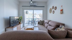 A modern living room with a sofa, a TV, a small table, and a balcony with a view of buildings. - Citrus 3-Bedroom Apartment in Larnaca (Larnaca)