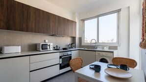 A modern kitchen with a microwave, stove, and sink, a dining area with a table and chairs, and a large window providing a view of the city. - Citrus 3-Bedroom Apartment in Larnaca (Larnaca)