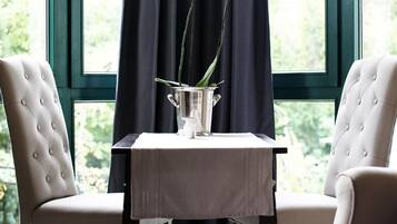 Een eettafel met een vaas witte bloemen, twee gestoffeerde stoelen en uitzicht op groen door een raam met donkere gordijnen.