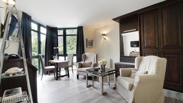 A living room with a sofa, armchairs, a coffee table, and a ladder. There is a window with curtains, a painting on the wall, and a television. - Höhen Hotel Viersen (Viersen)