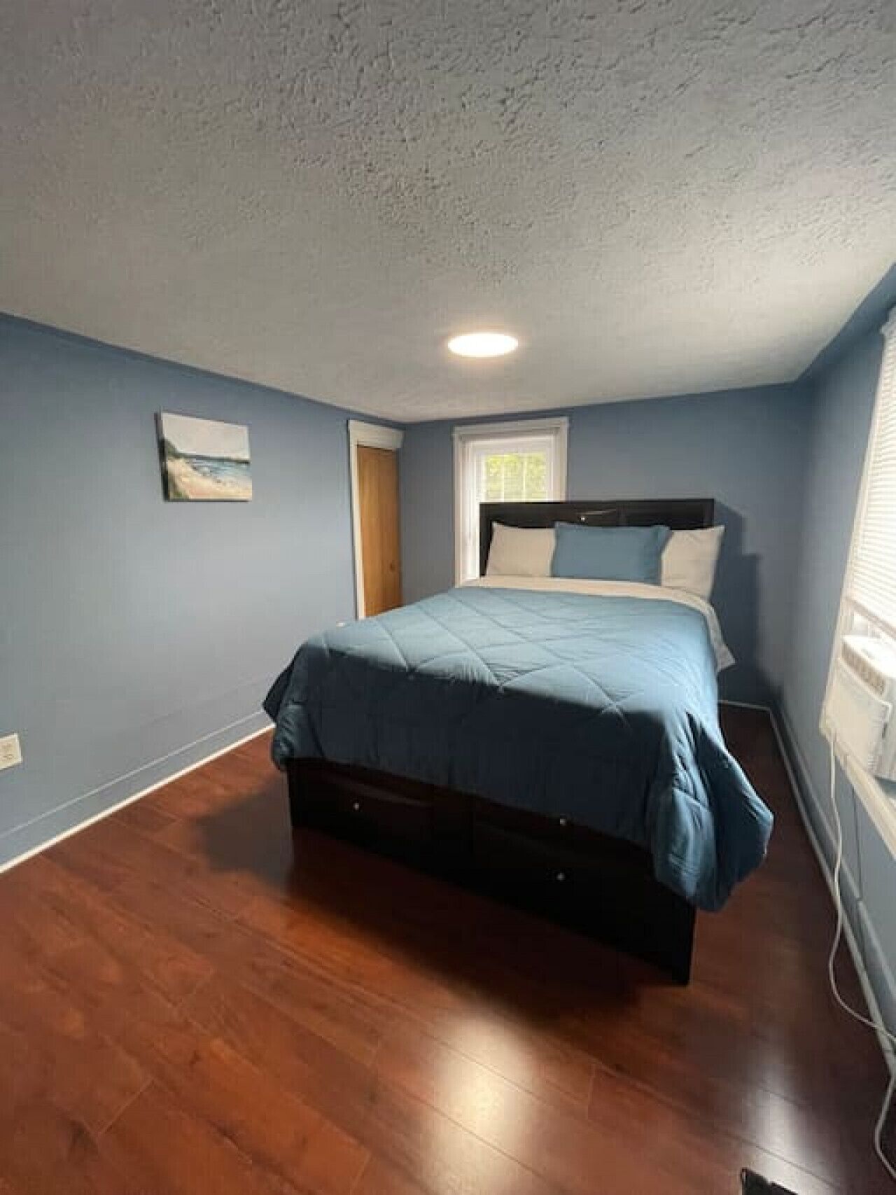 Een slaapkamer met een bed, een raam, een deur en een schilderij aan de muur.