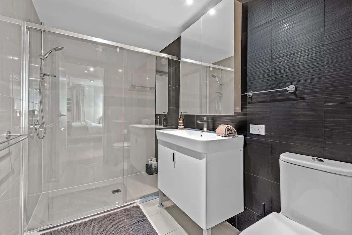 Un baño moderno con ducha de vidrio, tocador blanco y pared revestida de azulejos oscuros.