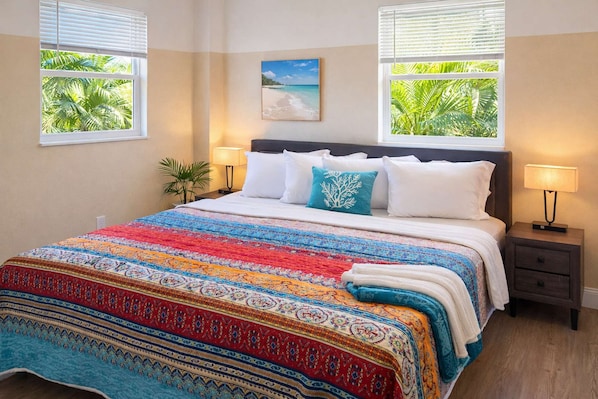 A bedroom with a bed, two bedside lamps, a nightstand, a painting, and windows with tropical views.