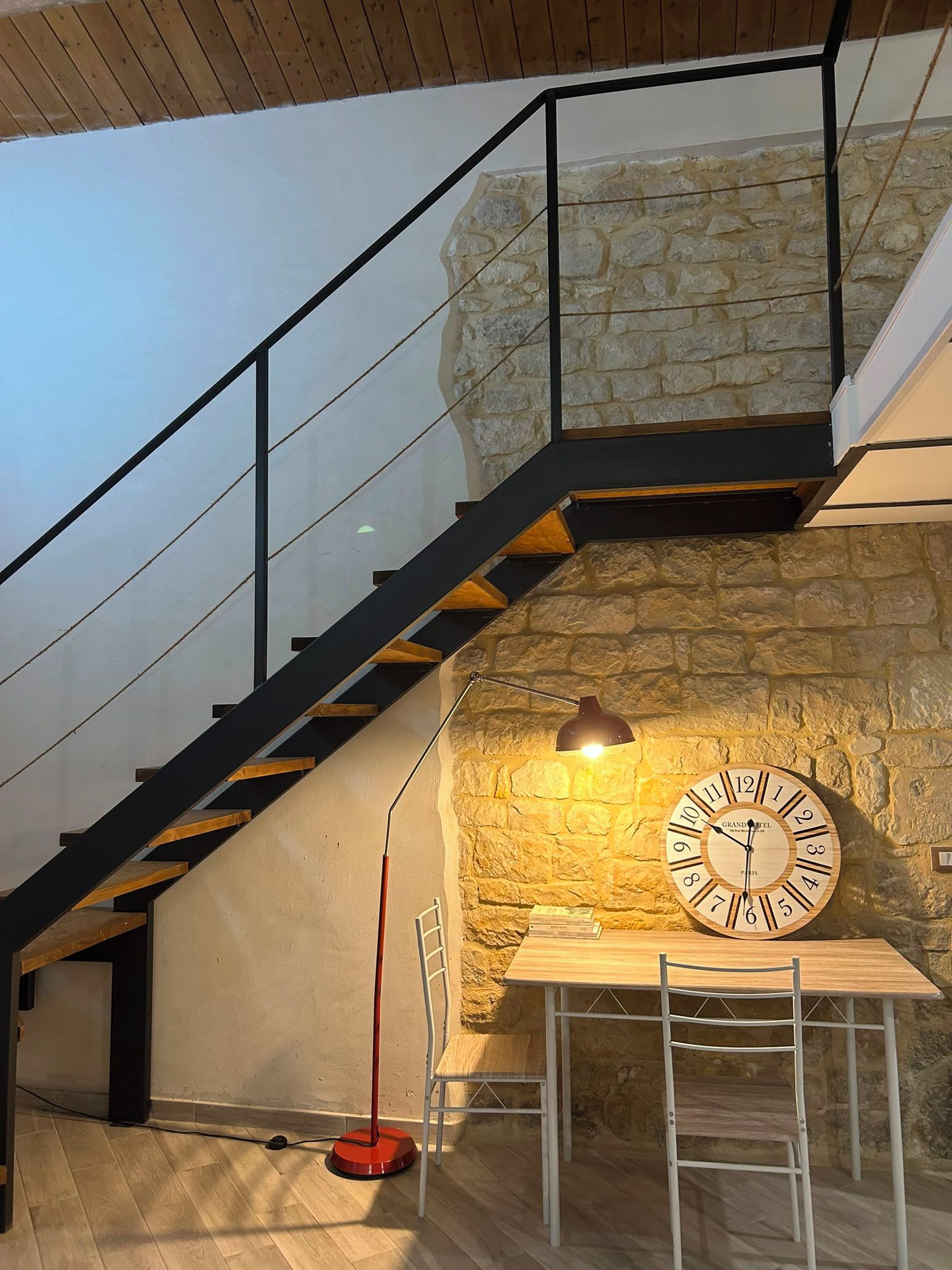 A staircase with a glass railing, a stone wall, a wooden table with chairs, and a wall clock.