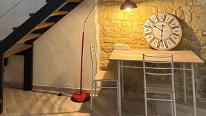 Una escalera con barandilla de cristal, un muro de piedra, una mesa de madera con sillas y un reloj de pared.