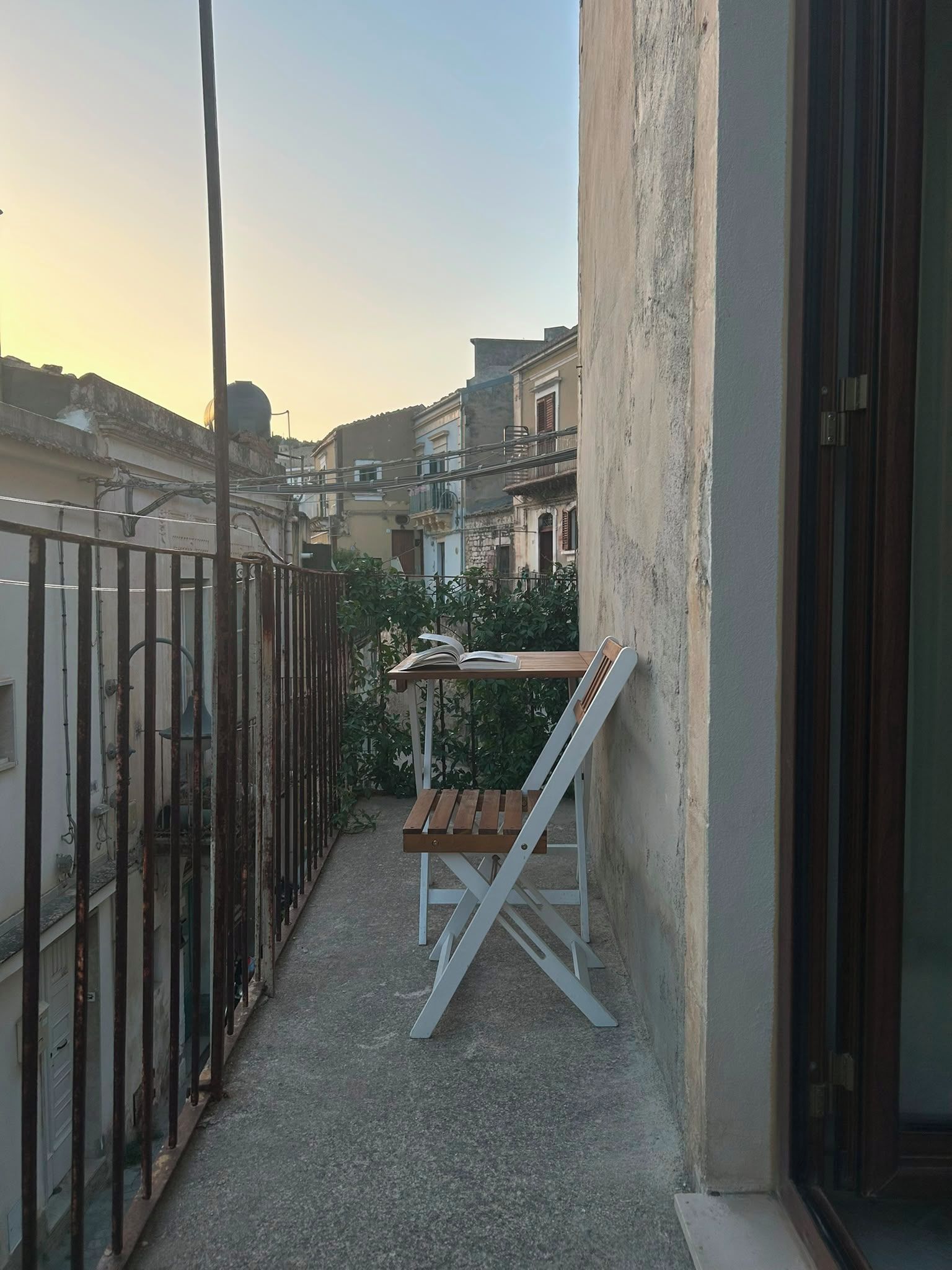 Un étroit balcon extérieur avec une table en bois et une chaise blanche, une clôture en métal et des bâtiments en arrière-plan.