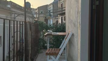 Un étroit balcon extérieur avec une table en bois et une chaise blanche, une clôture en métal et des bâtiments en arrière-plan.