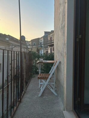 Un balcón exterior estrecho con una mesa de madera y una silla blanca, una cerca de metal y edificios al fondo.