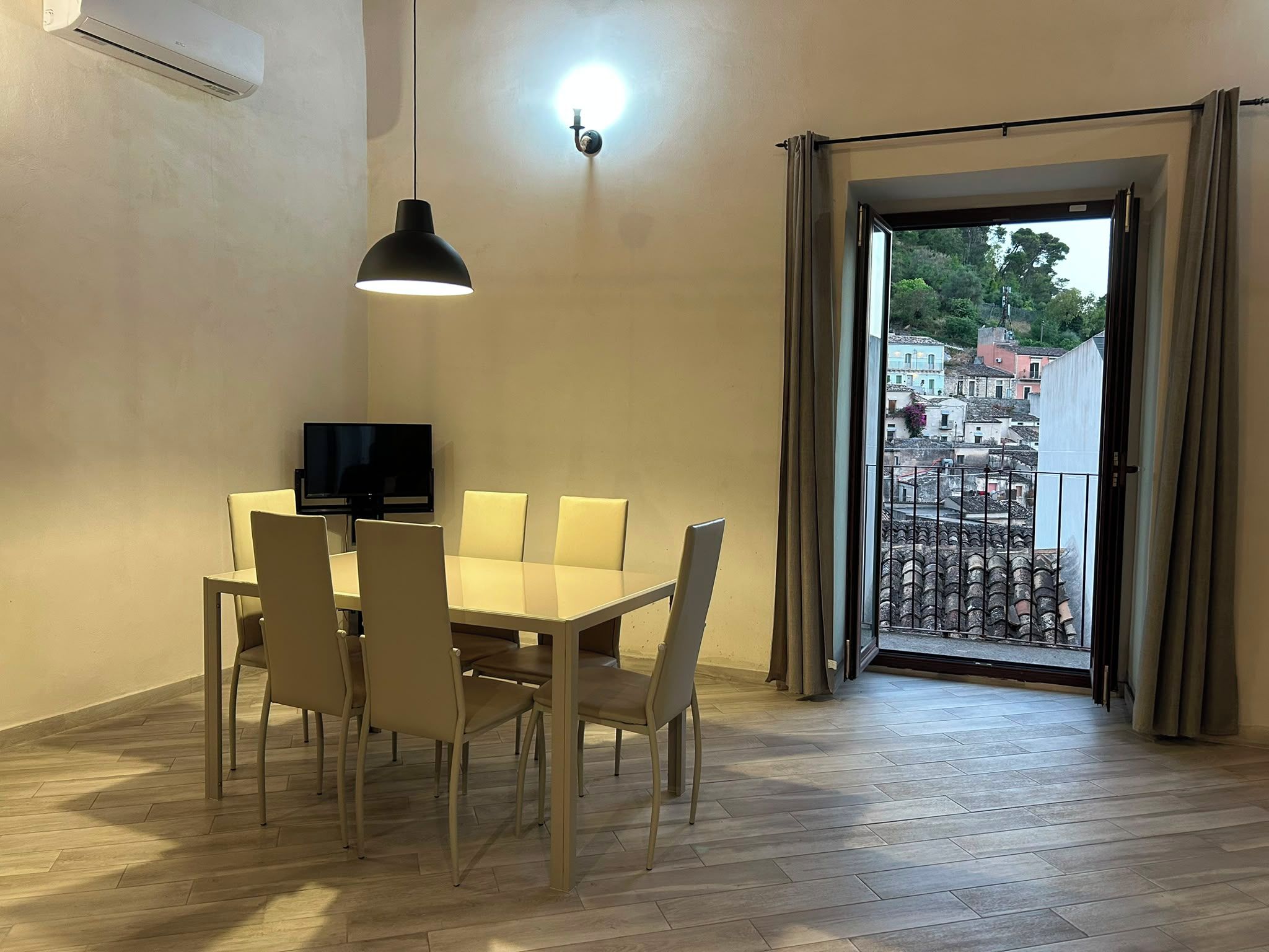 A dining area with a table and chairs, a television, and a view of a balcony with buildings.
