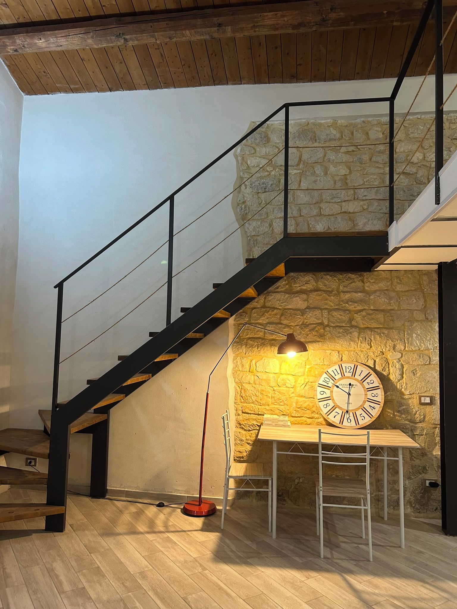 Una escalera con un muro de piedra, una mesa de madera con un reloj y una lámpara roja en el suelo.