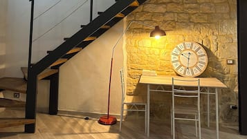 Una escalera con un muro de piedra, una mesa de madera con un reloj y una lámpara roja en el suelo.