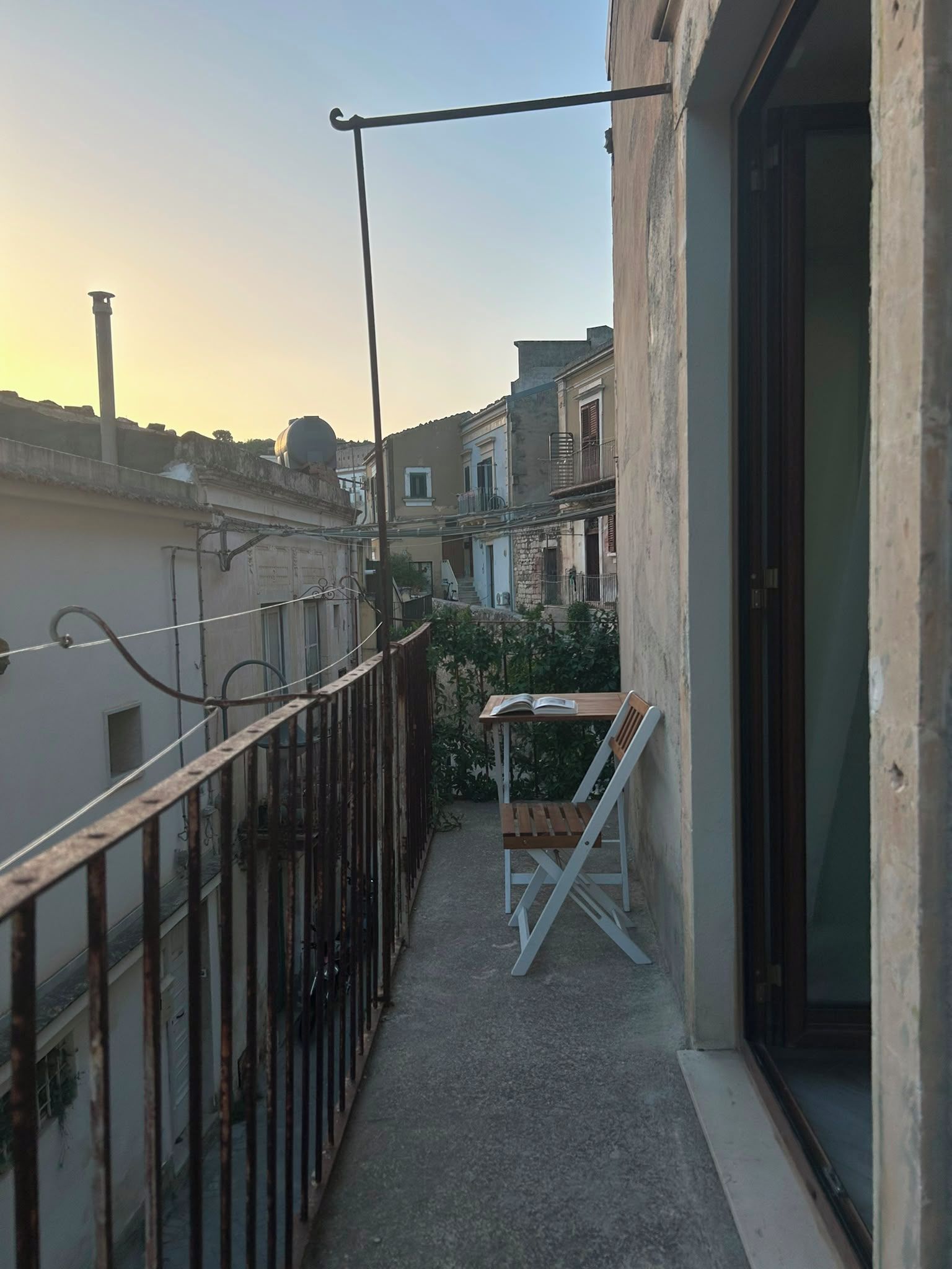 A narrow balcony with a metal railing, a small table with a chair, and a view of adjacent buildings.