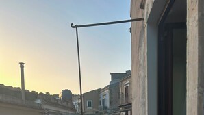 Un balcón estrecho con barandilla de metal, una mesa pequeña con una silla y vistas a edificios cercanos.