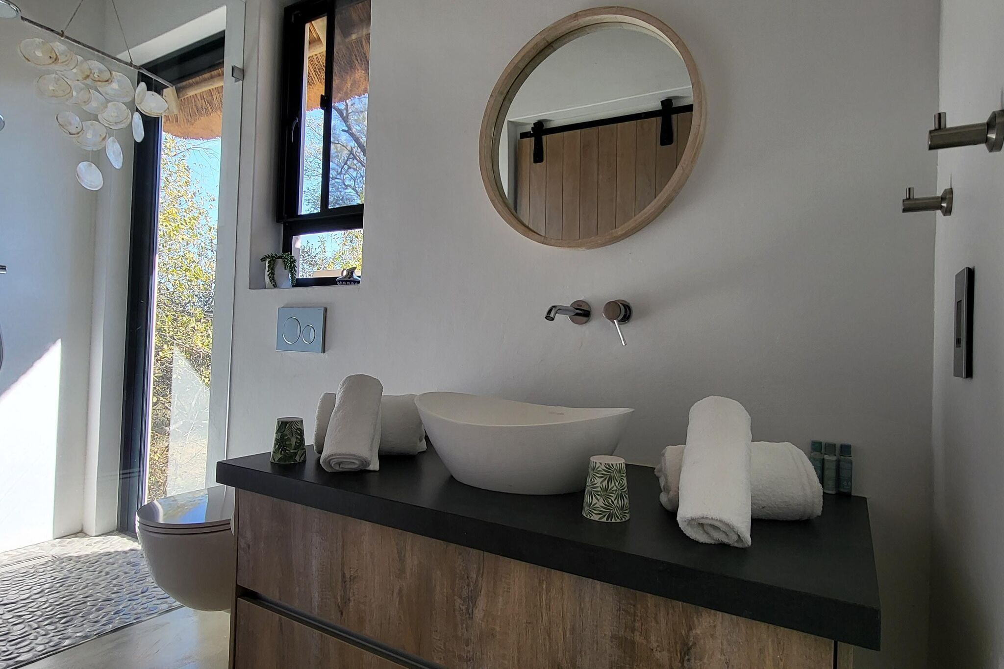 A modern bathroom with a large sink, a round mirror, and a window with a view of trees.