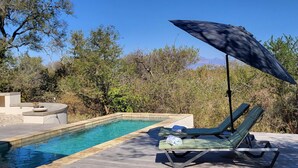 Un área de piscina con sillones de descanso y una sombrilla, rodeada de árboles y un cielo despejado.