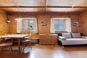 A wooden cabin interior with a dining table, chairs, a sofa, and two windows with curtains.