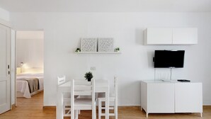 Una habitación moderna y minimalista con una mesa y sillas blancas para comer, un mueble blanco con un televisor de pantalla plana y una cama al fondo.