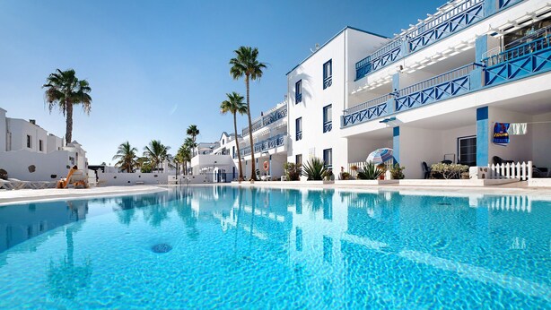 Una piscina con agua azul cristalina, edificios blancos con balcones azules y palmeras.
