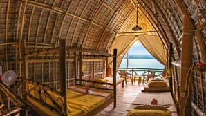 Een kamer met rieten dak, een bed, een ventilator en uitzicht op zee.