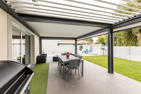 A covered patio with a dining table and chairs, a grill, and a view of a swimming pool and garden.