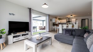 A modern living room with a flat-screen TV, grey sofa, and a dining area with a kitchen in the background. - Villa "Bassin d'Arcachon" with Private Pool, Private Terraces and Wi-Fi (ARES)