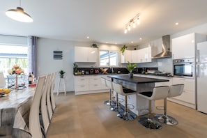 A modern kitchen with white cabinets, a central island with bar stools, stainless steel appliances, and a large window. - Holiday Home "Villa Bassin D'Arcachon" with Private Pool, Private Terraces and Wi-Fi (Ares)