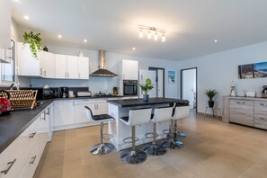 A modern kitchen with white cabinets, a central island with bar stools, a stainless steel range hood, and a large window. - Holiday Home "Villa Bassin D'Arcachon" with Private Pool, Private Terraces and Wi-Fi (Ares)
