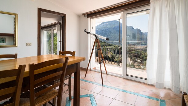 A dining area with a wooden table and chairs, a telescope, and a large window with a mountain view. - Holiday Apartment "Casa Su Craminu" with Mountain View, Balcony & Wi-Fi (Dorgali)
