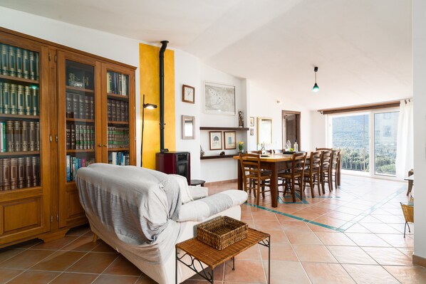 A living room with a sofa, dining table, chairs, a bookshelf, and a view through a large window.
