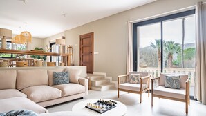 A spacious living room with a large window, a sofa, two armchairs, a coffee table with a chess set, and a staircase leading to an upper floor. - Villa "Son Simo D'Alt" with Mountain View, Private Pool & Wi-Fi (Crestatx)