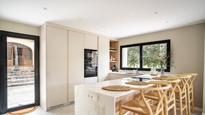 A modern kitchen with a marble island, wooden chairs, and a large window. - Villa "Son Simo D'Alt" with Mountain View, Private Pool & Wi-Fi (Crestatx)