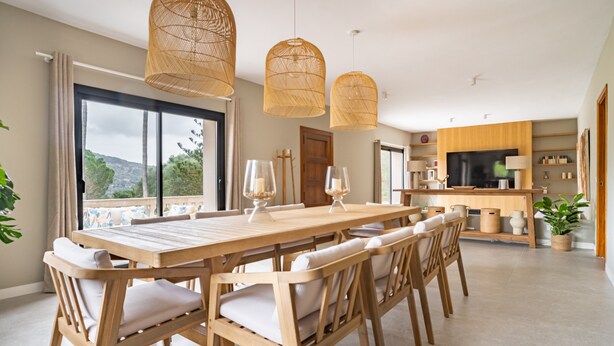 A dining area with a wooden table and chairs, a large window with curtains, and a kitchen area with wooden cabinets and a TV. - Villa "Son Simo D'Alt" with Mountain View, Private Pool & Wi-Fi (Crestatx)