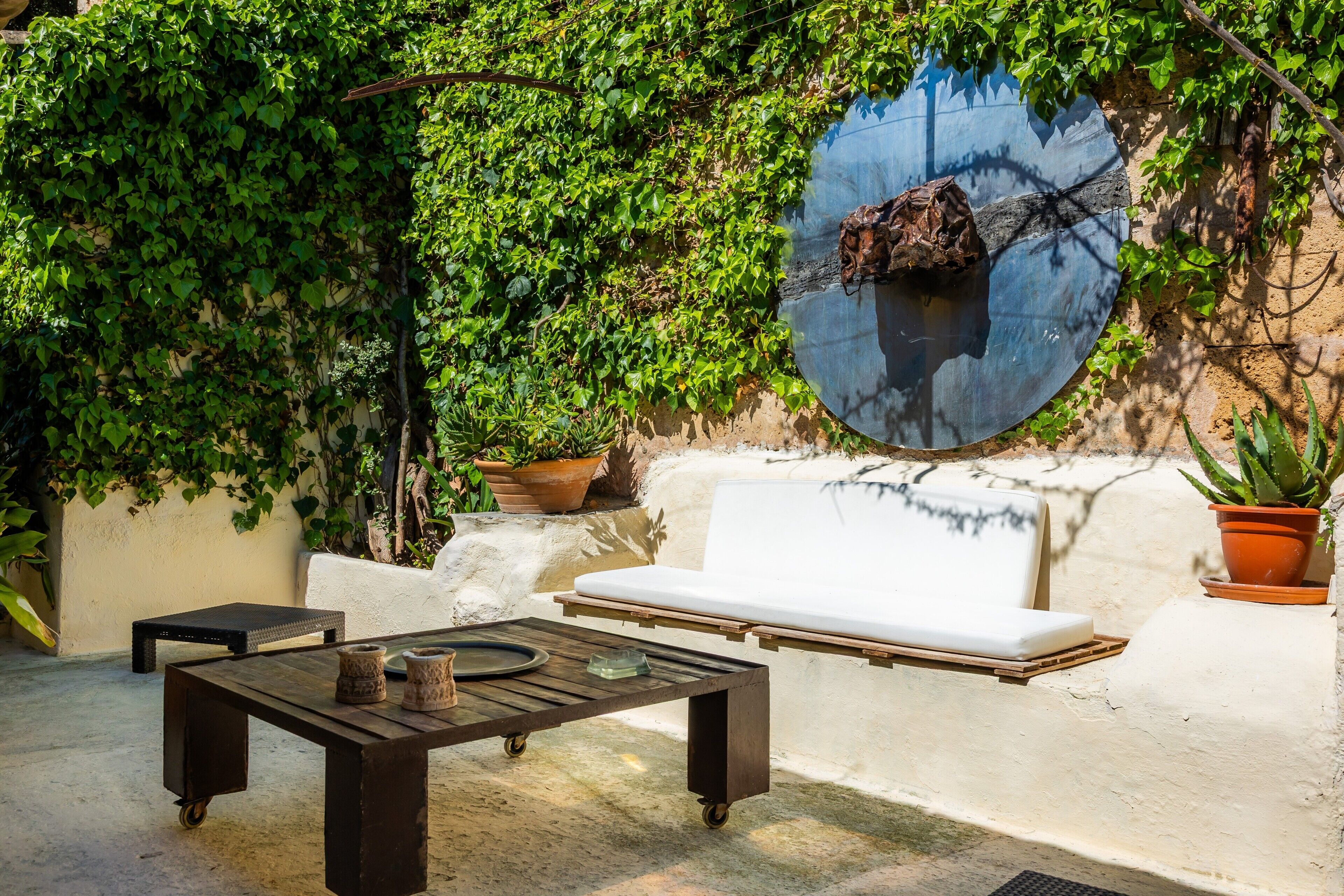 An outdoor patio with a round mirror, a white sofa, a wooden table, and potted plants.