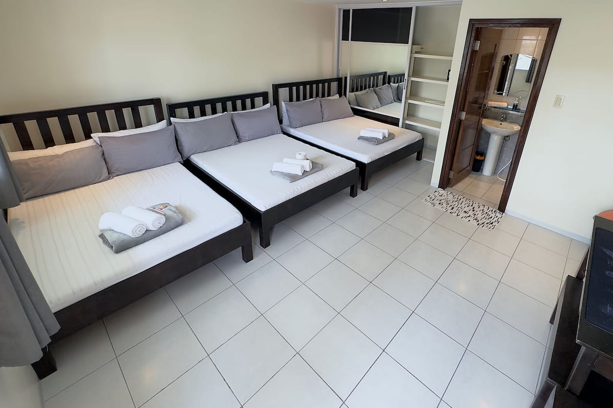 A hotel room with three single beds, a TV, a bathroom with a sink, and a mirror.