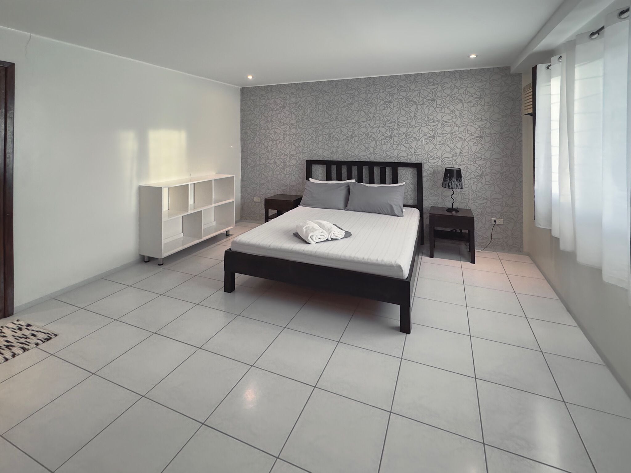 A modern bedroom with a large bed, a white wardrobe, a bedside table with a lamp, and a patterned wall.