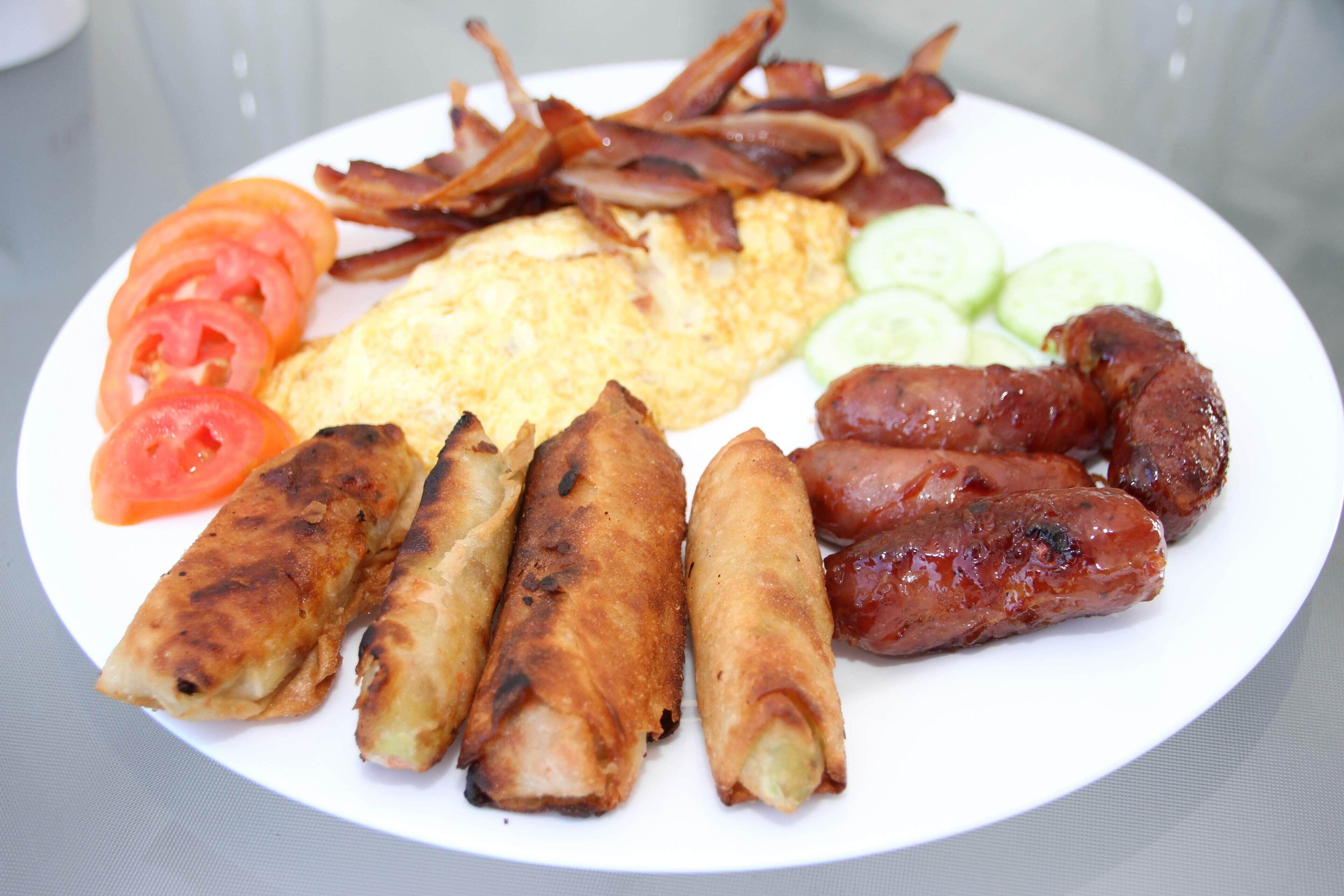A plate of food including eggs, bacon, sausages, spring rolls, and sliced tomatoes and cucumbers.