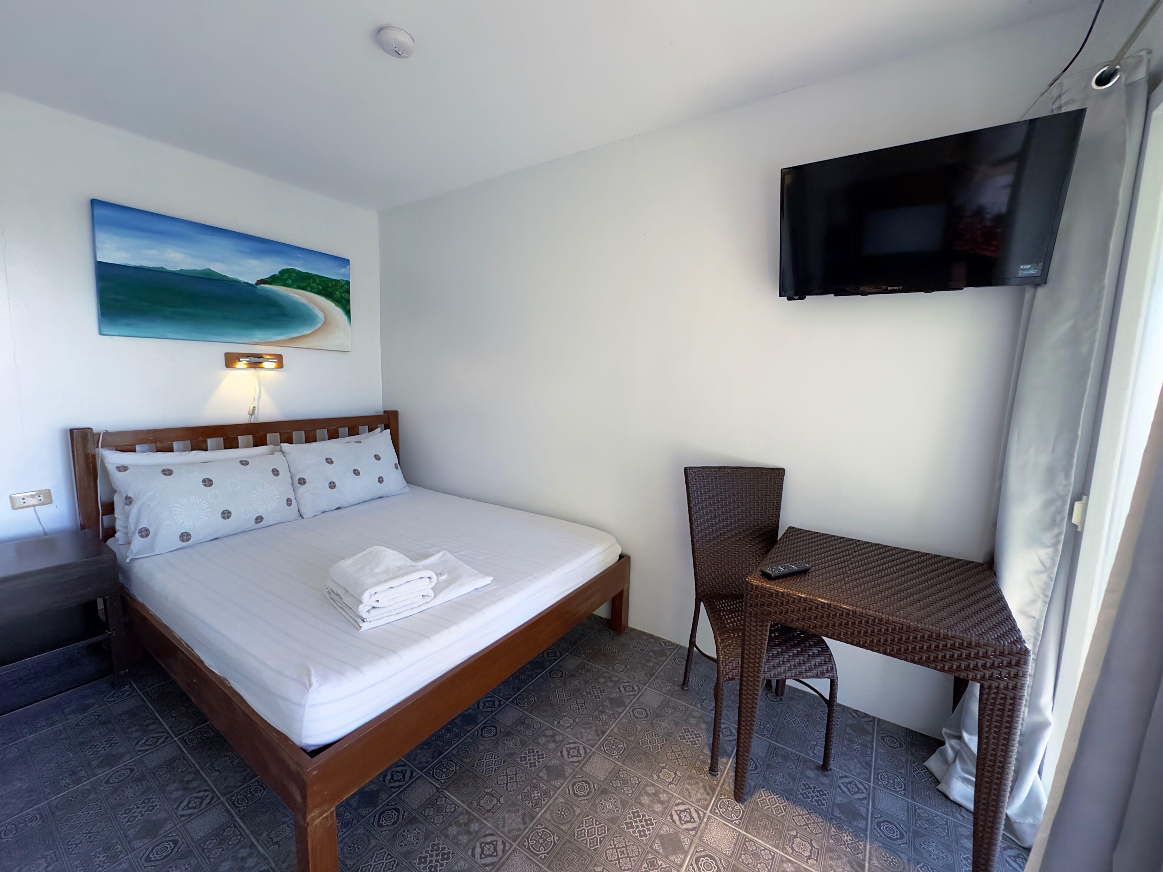 A hotel room with a bed, a desk, a TV, and a painting of a beach.