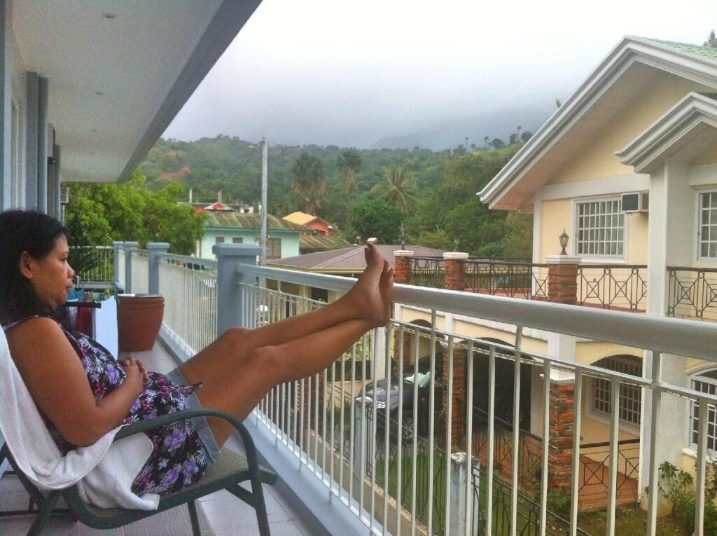 Un balcón con una persona sentada, con vistas a una zona residencial con casas y vegetación.