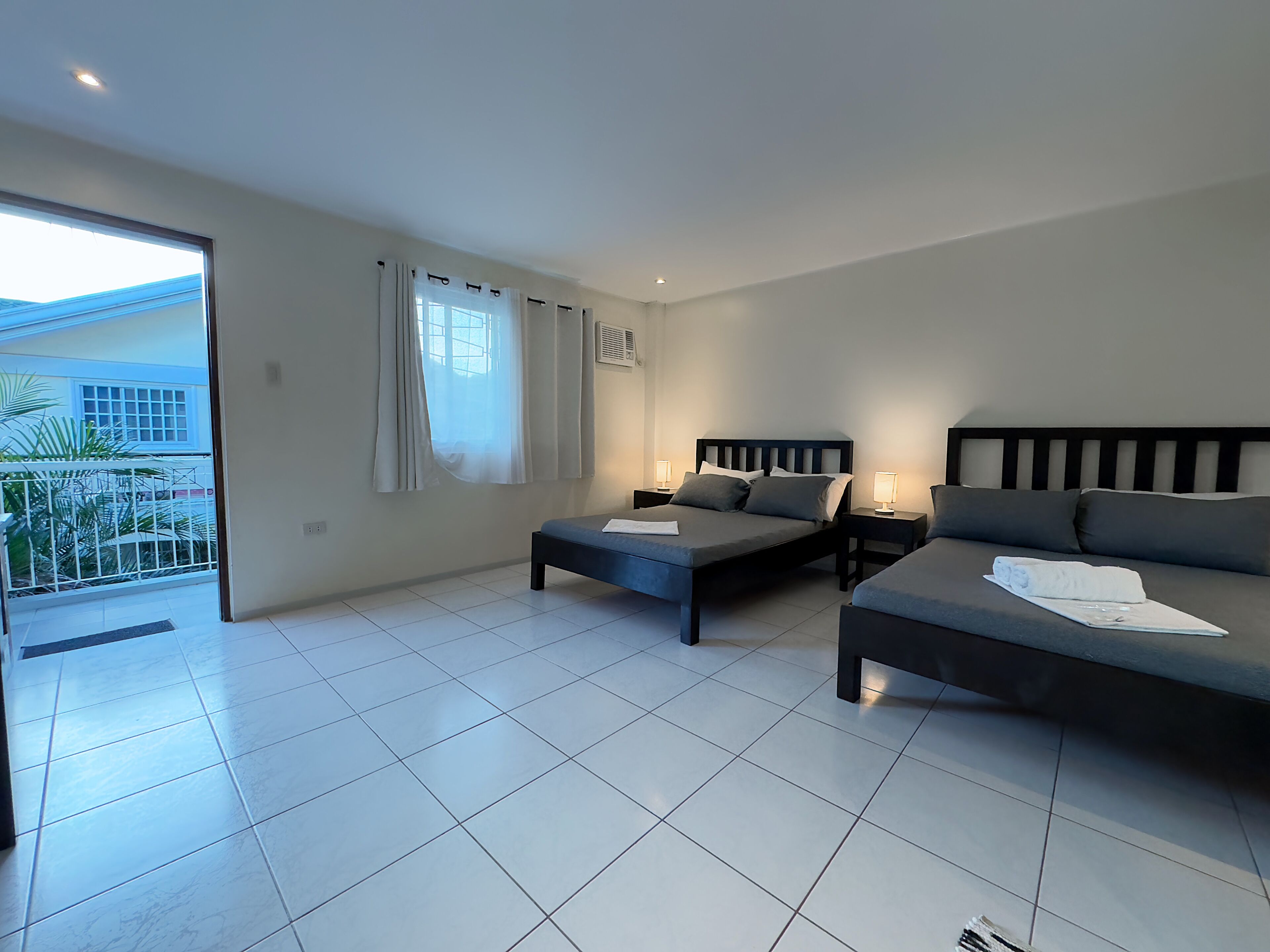 A modern bedroom with two single beds, a sofa, and a balcony with a view of a house and greenery.