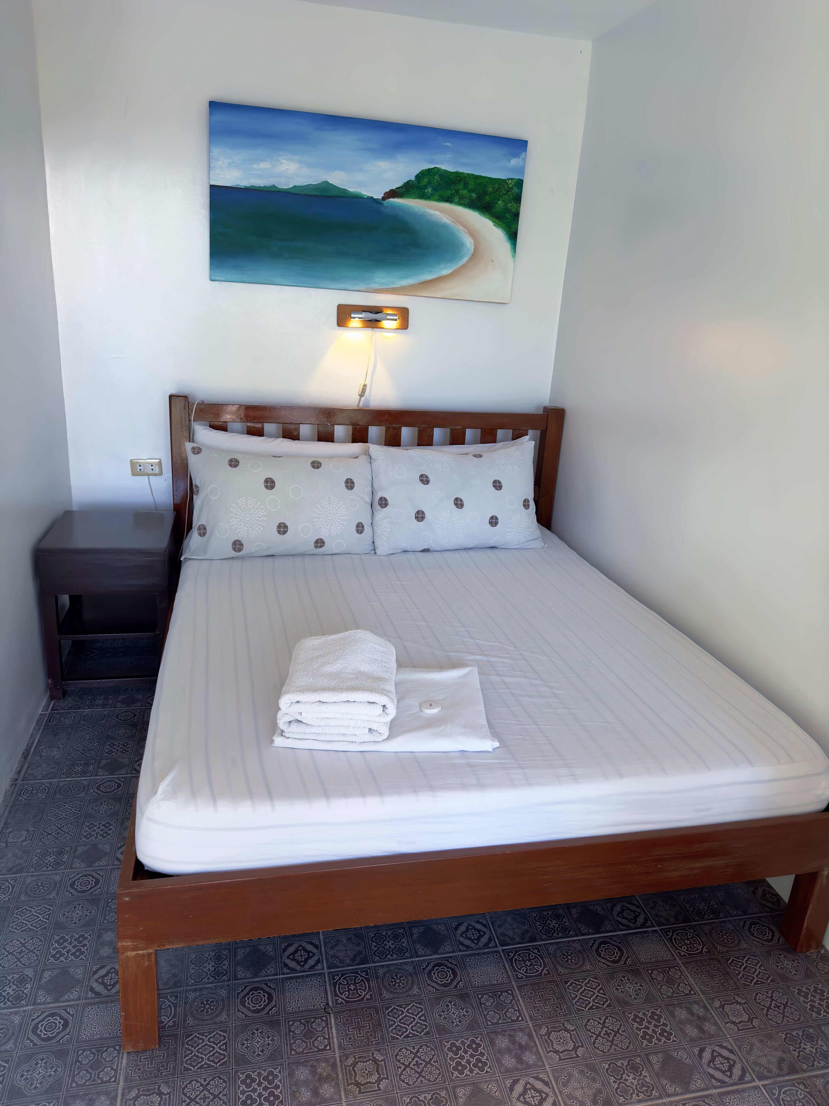 A neatly made bed with a white comforter and pillows, a wooden headboard, a nightstand with a lamp, and a painting of a beach scene on the wall.