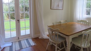 Ein Esszimmer mit einem Holztisch und weißen Stühlen, einer Tür, die in einen Garten führt, und einem gerahmten Bild an der Wand.