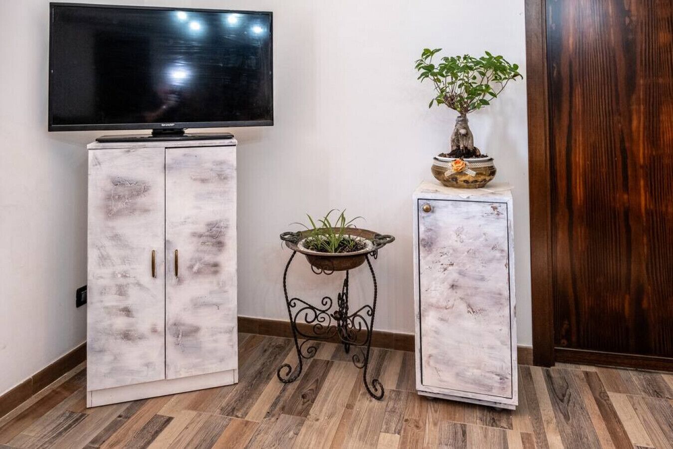 Une pièce avec un téléviseur fixé au mur, deux armoires au fini vieilli, une petite table sur laquelle est posée une plante en pot et un plancher en bois.