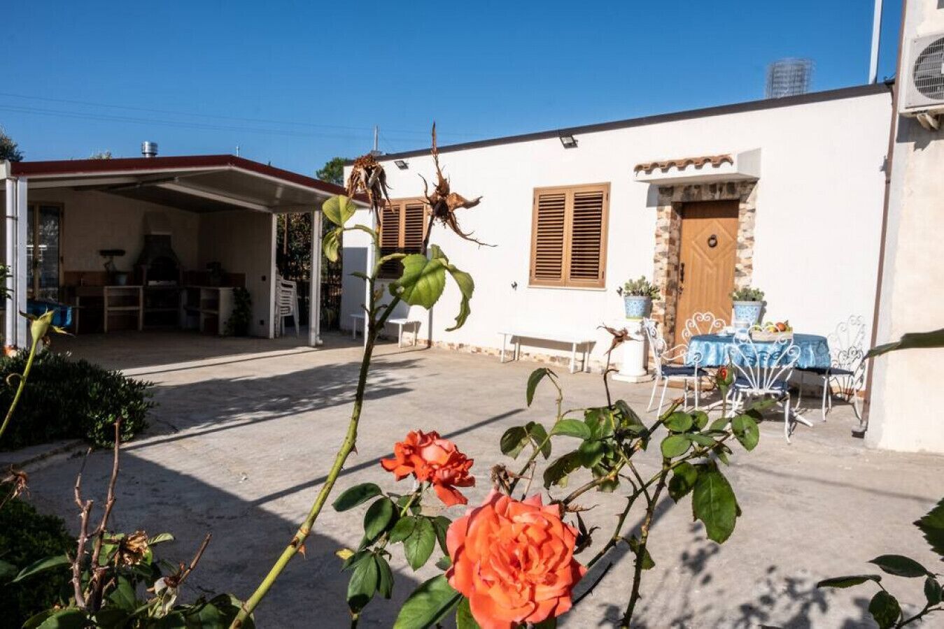 Un edificio blanco con un porche cubierto, una mesa con mantel azul y un rosal al frente.