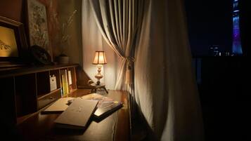 A dimly lit room with a desk, a lamp, and a view of a cityscape at night.