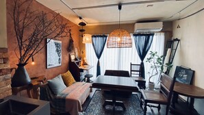 A cozy room with a bed, a dining table, and chairs. There is a large window with curtains, a hanging light fixture, and a potted plant. - TOE LIBRARY (Tokyo)