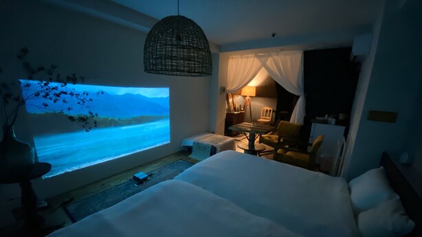 A hotel room with a large bed, a small seating area, a TV screen displaying a scenic view, and a hanging light fixture. - TOE LIBRARY (Tokyo)